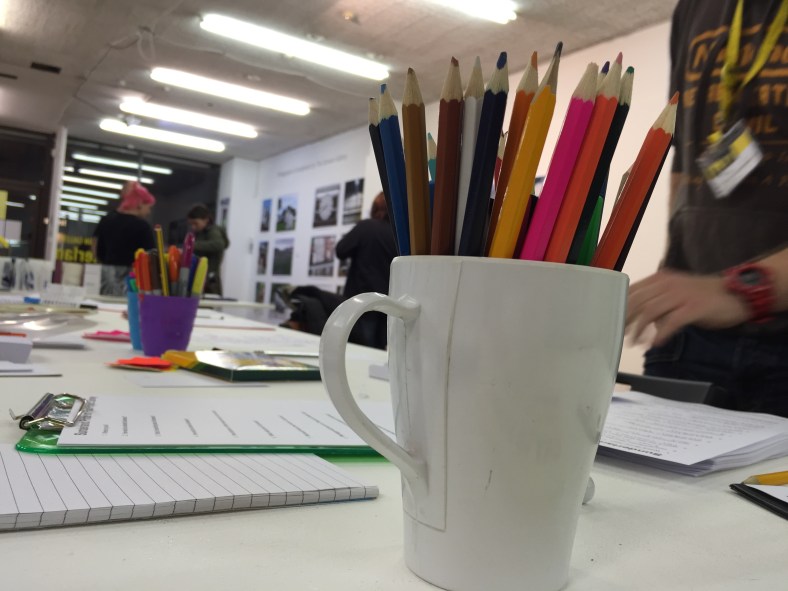 Some coloured pencils and surveys on the table at the Caravan Gallery's open event. In the background, some people are chatting and looking at the photography on display