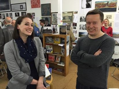 Two attendees to the Caravan Gallery's event . Behind; a small crowd of people, a bookshelf containing several books with their fronts on display, and a wall of photographs and artwork.